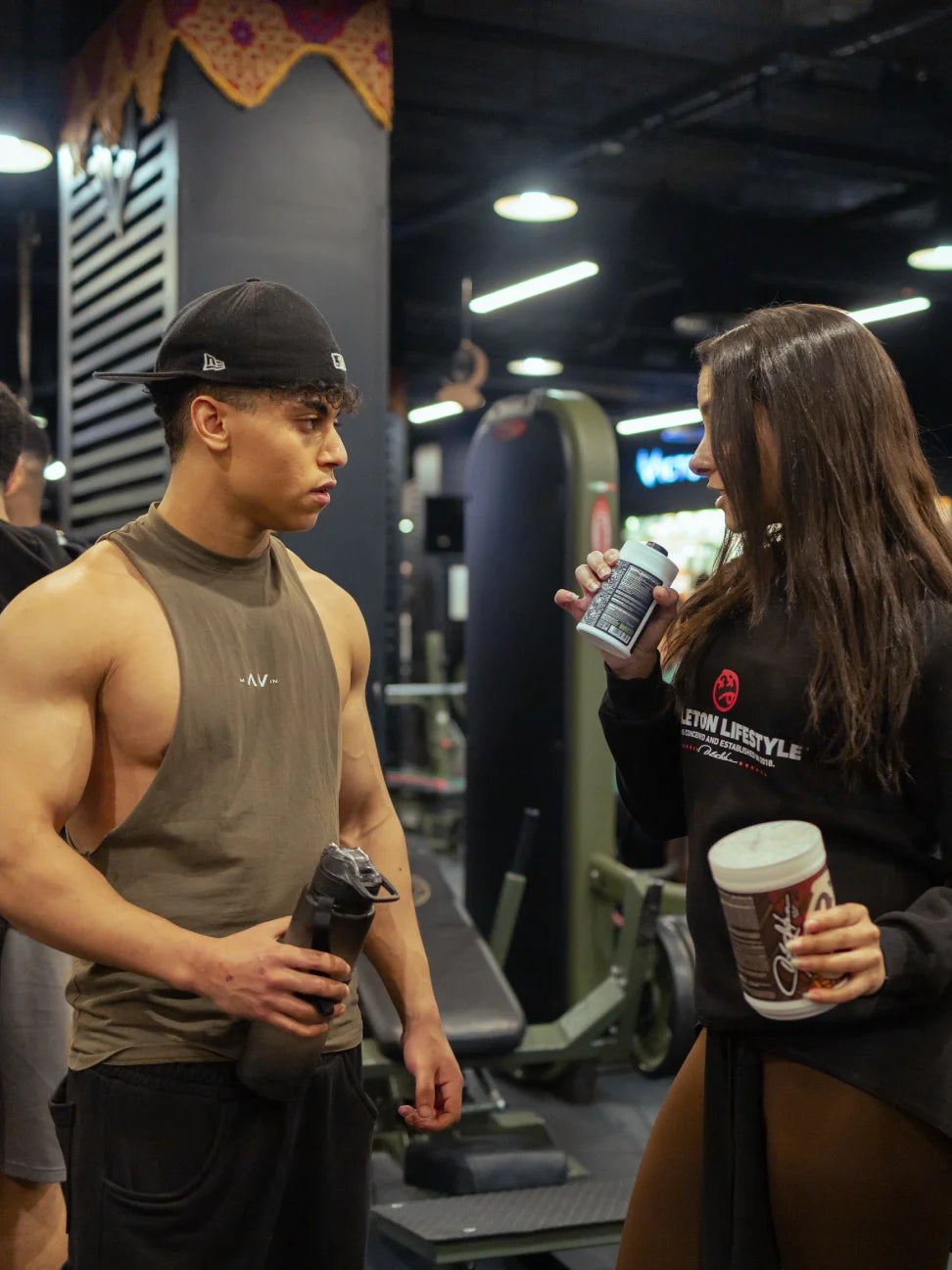 Two people in a gym setting, one holding a skeleton spooky jar and the other a skeleton protein shake.